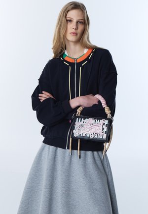 Jonge vrouw met lang blond haar draagt een zwart jack met rits en een grijze rok, en houdt een zwarte handtas met roze bloemenborduurwerk en touwgrepen vast.