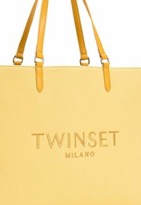 Rumena torba z dvema ročajema, izdelana iz teksturiranega materiala. Ima embosiran logotip "TWINSET MILANO" v usklajeni rumeni nitki.