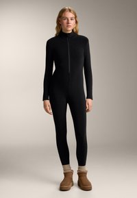 Čierny body s celým zipsom, dlhými rukávmi, priliehavým strihom, hladkou textúrou a vysokým golierom, doplnený hnedými členkovými topánkami a ponožkami.