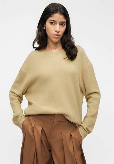 Pull en maille côtelée beige avec un col rond et des épaules tombantes, associé à un pantalon plissé marron taille haute.