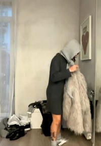 Inimene halli kapuutsiga pusa ja kleitidega hoiab suurt kohevamat mantlit, seisab segases toas raamitud südamekunstiteose lähedal.