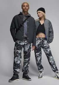 Två modeller bär camo-mönstrade byxor i grått och svart. En modell har en svart jacka och en grå hoodie; den andra bär en kort svart topp och en bomberjacka.