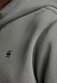 Sudadera con capucha gris hecha de una tela suave. Presenta un logo en negro, costuras visibles y un ajuste cómodo alrededor del cuello y los hombros.