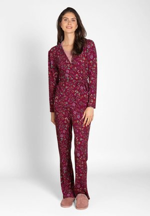 Woman wearing a maroon floral wrap top and matching pants with pink slippers, standing against a plain white background.