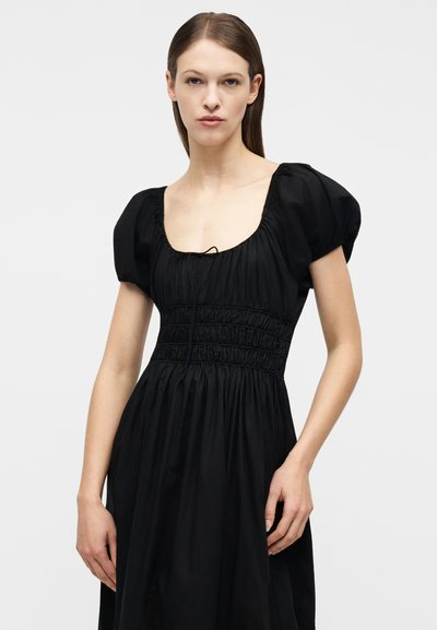 Γυναίκα με ίσια καστανά μαλλιά που φοράει μαύρο φόρεμα με φουσκωτά μανίκια και ραμμένη μέση, στέκεται μπροστά σε απλό λευκό φόντο.