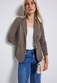 Blazer en tricot marron au design ajusté, avec deux poches avant, associé à un haut blanc et un jean bleu, tenant une pochette en osier beige.