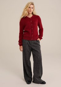 Sarkans, teksturēts kardigans ar apaļu dekoltē un pogu aizdari, apvienots ar platām tumšām biksēm ar rakstu un melniem apaviem.