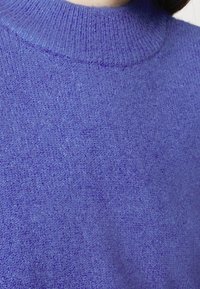 Pull tricoté violet avec un col côtelé haut, une texture douce et une coupe légèrement ample. Le tissu présente un léger éclat et un motif uniforme.