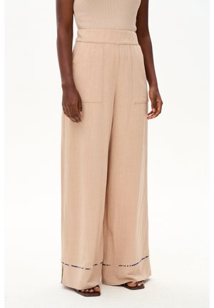 Kobieta nosząca beżowe, szerokie spodnie z wysokim stanem, naszywanymi kieszeniami i rozcięciami u dołu, zestawione z brązowymi sandałami z odkrytymi palcami.