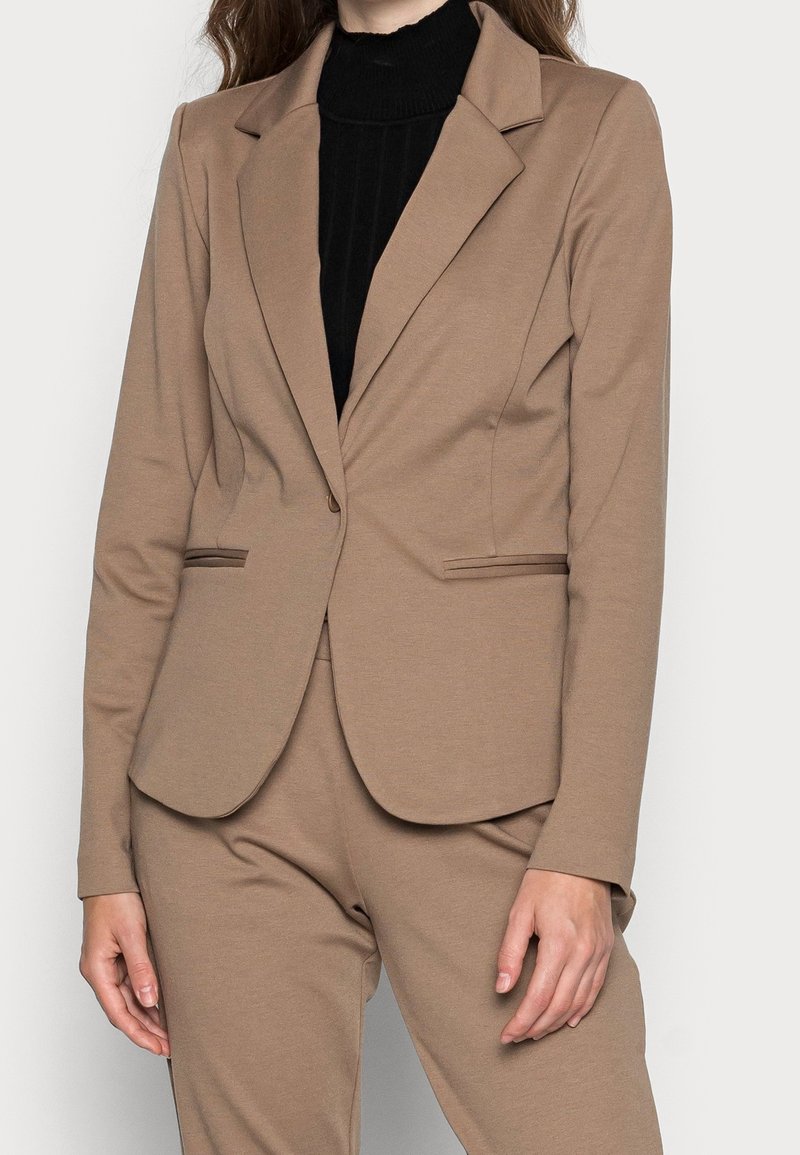 Béžový blazer vyrobený z elastického materiálu, s klopovým límcem, dvěma předními kapsami s jemným lemováním a přiléhavým střihem.