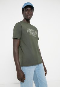 Khaki zielona bawełniana koszulka z krótkim rękawem, z trójkątnym logo "Guess" w jasnym kolorze. Noszona z jasnoniebieskimi dżinsami.