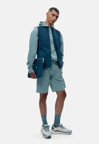 Šviesiai mėlynas džemperis ir šortai derinami su tamsiai mėlyna siūta liemene. Jis laiko tamsiai mėlyną nešiojamojo kompiuterio dėklą, užsidėj ęs šviesius sportinius batelius ir mėlynas kojines.