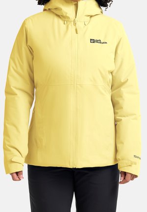 Persona que lleva una chaqueta con capucha amarilla Jack Wolfskin con cremallera frontal y puños ajustables, combinada con pantalones negros.