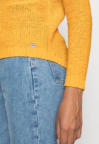 Pull en maille jaune avec un design à trames ouvertes et texturées, associé à un jean en denim bleu clair. Il possède un petit tag logo "ONLY" au bas.