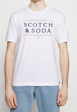 Mężczyzna w białej koszulce z krótkim rękawem z czarnym napisem „Scotch & Soda” oraz jasnoniebieskich dżinsach, stojący na jednolitym tle.