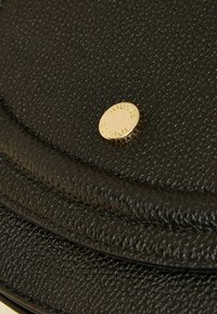 Čierna kožená taška s textúrovaným povrchom, zaobleným tvarom a zlatým emblémom s nápisom "JOUTE." Hladký povrch s viditeľnými švami.