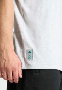 Harmaa lyhythihainen t-paita, jossa on teksturoitu pinta, vihreä Adidas-logo ja Real Madridin tunnus alahelmassa.