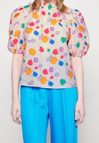 Bloemige blouse in een lichte stof, met een textuur in crème als achtergrond en kleurrijke bloemen in oranje, roze, paars en groen. Tulp-schoenen.