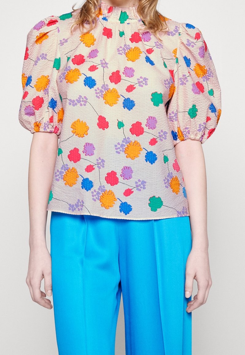 Bloemige blouse in een lichte stof, met een textuur in crème als achtergrond en kleurrijke bloemen in oranje, roze, paars en groen. Tulp-schoenen.