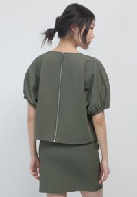 Top verde oliva con maniche a sbuffo, design corto e cerniera centrale sul retro. Tessuto liscio con una silhouette moderna.