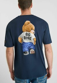 Man in een marineblauw t-shirt met een teddybeerafbeelding op de rug, de beer draagt een wit shirt met het label "BIG BOSS" en blauwe jeans.