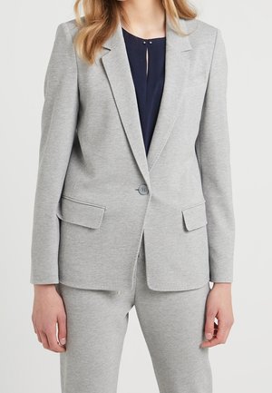 Frau trägt einen hellgrauen Blazer mit einem Knopf und Pattentaschen, kombiniert mit passenden Hosen und einer dunkelblauen Bluse darunter.