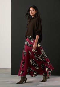 Bruine oversized trui gecombineerd met een lange bordeauxrode rok met zwarte bloemenpatronen, aangevuld met bruine haklaarzen tot de enkel.