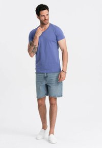 T-shirt di cotone viola con scollo a V, abbinato a pantaloni corti di jeans blu chiaro. Il modello indossa scarpe slip-on bianche e ha un tatuaggio sul braccio sinistro.