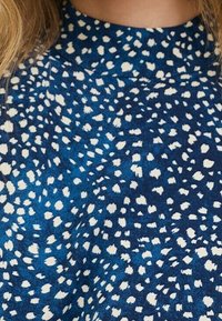 Tissu bleu avec un motif à pois blancs. Encolure ronde avec une texture subtile. Matériau doux qui tombe doucement. Pas de matériel ou d'accents visibles.