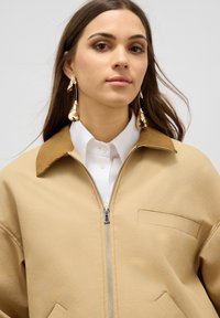 Beige ritsjack met een bruine kraag, voorzien van een zijzak en een witte shirt eronder. Model draagt glanzende hangende oorbellen.