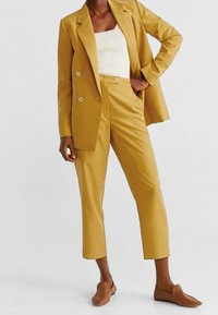 Veste jaune moutarde et pantalon court assorti en tissu lisse, associés à un haut blanc côtelé et des chaussures marron à enfiler.