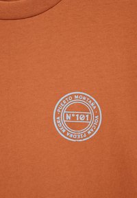 Tissu orange rouille avec un petit tampon circulaire blanc portant l'inscription « No 101 Puerto Montaña Volcán Piedra Negra » près du col.
