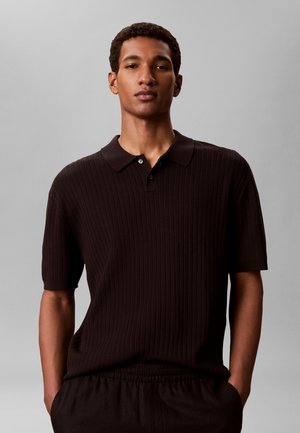 Młody mężczyzna z krótkimi kręconymi włosami, ubrany w czarną prążkowaną koszulkę polo z krótkim rękawem i czarne spodnie na neutralnym tle.