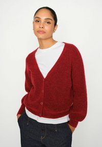 Cardigan roșu din lână cu decolteu adânc în V, închidere cu nasturi și manșete cu nervuri. Purta peste un tricou alb și asortat cu blugi albastru închis.