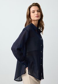 Navy sheer button-up shirt with long sleeves, light fabric, a collar, and metallic buttons. Features a curved hem and relaxed fit.