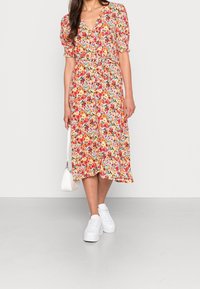 Donna che indossa un vestito midi floreale colorato con maniche a sbuffo, sneakers bianche e tiene una piccola borsa bianca.