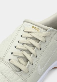 Μπεζ αθλητικό παπούτσι με υφή, επίπεδα κορδόνια στο ίδιο χρώμα και διακριτικό λευκό λογότυπο Nike στο πλάι.