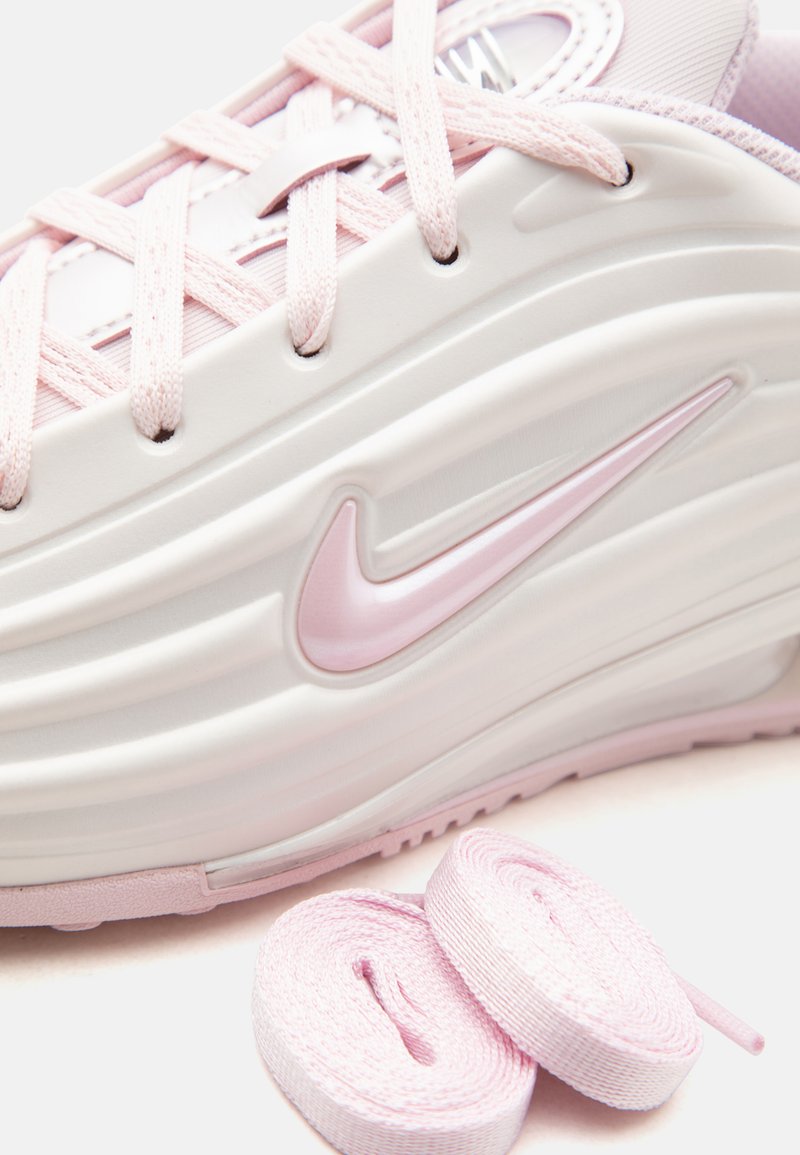 Primer plano de una zapatilla Nike rosa claro y blanca con los cordones enrollados a juego junto a ella sobre una superficie blanca.