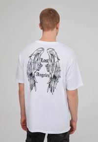T-shirt bianca in cotone a maniche corte con grandi grafiche di ali nere e testo "Lost Angeles" sul retro. Design semplice con scollatura a girocollo.