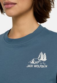 T-shirt in cotone blu con un'immagine bianca di un campeggio con alberi e una tenda, con il marchio "JACK WOLFSKIN" sotto il design.