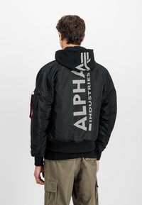 Zwarte bomberjack met zilveren "ALPHA INDUSTRIES" logo op de achterkant, gemaakt van nylon, met ribgebreide manchetten en een capuchon. Voorzien van een ritszak aan de zijkant.