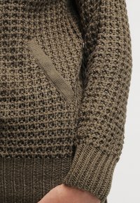 Suéter de punto en marrón tierra con una textura de tejido abierto, que presenta puños acanalados y un sutil bolsillo lateral.