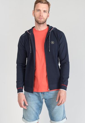Sweat à capuche marine avec un tissu texturé, accents blancs et rouges, fermeture éclair et détail de logo, porté sur une chemise orange et un short en denim.