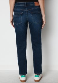 Jeans de denim azul oscuro con corte ajustado. Presenta dos bolsillos traseros y una etiqueta de cuero marrón en la cintura. Combinado con zapatillas blancas.