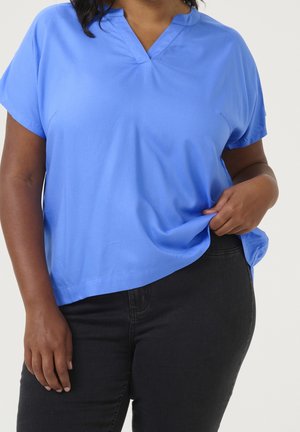 Persoon draagt een felblauw kortermig shirt en een zwarte broek, houdt het shirt lichtjes vast met één hand op heuphoogte.