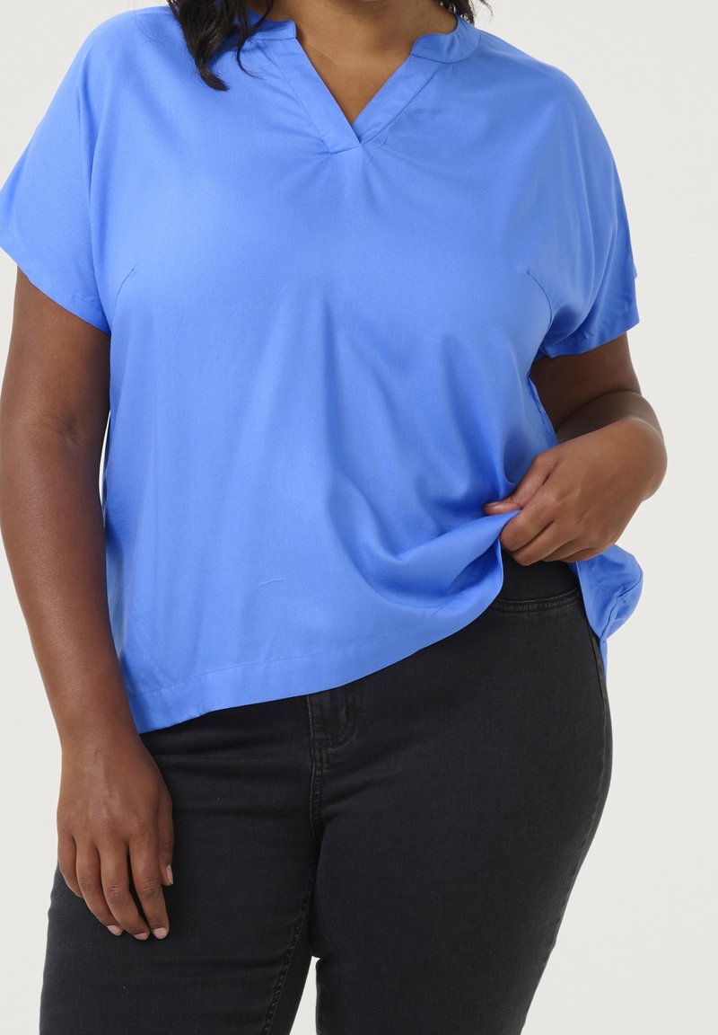 Personne portant une chemise bleu vif à manches courtes et un pantalon noir, tenant légèrement la chemise d’une main au niveau de la taille.
