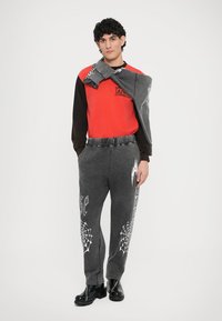 Pantalones deportivos grises con patrones de telaraña y texto blanco, combinados con una camisa de manga larga roja y negra. El modelo lleva botas negras.