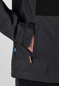 Main dans la poche latérale d'une veste sombre avec une sangle Velcro au poignet et une petite étiquette avec le drapeau suédois près de la poche.