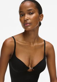 Juodas liemenėlės viršus su plonais reguliuojamais dirželiais, turintis išpjovą priekyje ir subtiliais sidabriniais akcentais. Lygus paviršius.