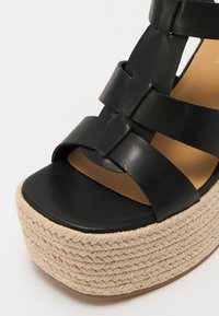 Črne usnjene sandale z wedges peto in pletenim jute podplatom. Imajo široke trakove in odprto obliko prstov, kar poudarja teksturiran podplat in naravne akcentke.